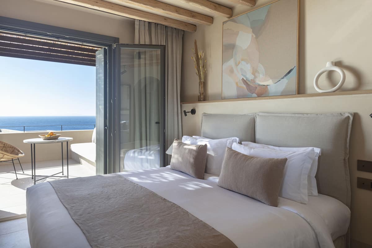 A bedroom with a large bed, a view of the ocean, a small table with a bowl of fruit, and a chair.