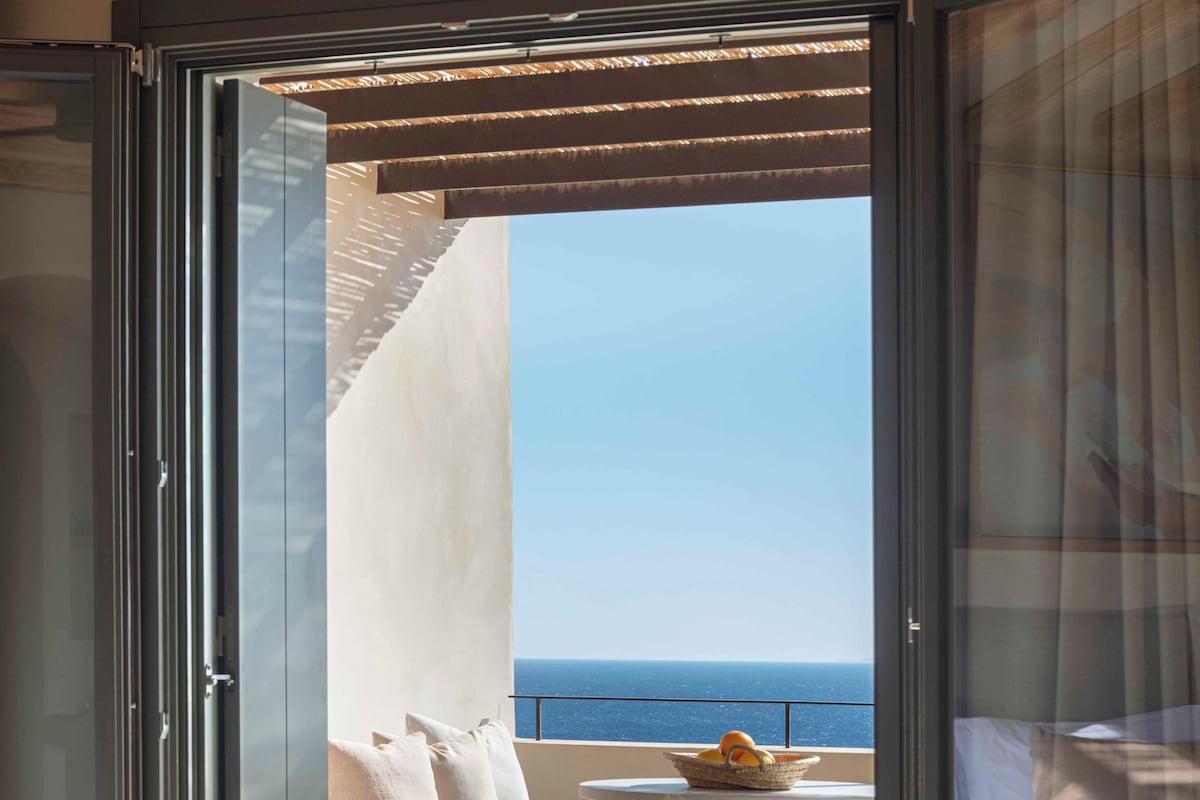 Een slaapkamer met een bed, een balkon met een tafel en een schaal fruit, en uitzicht op zee.