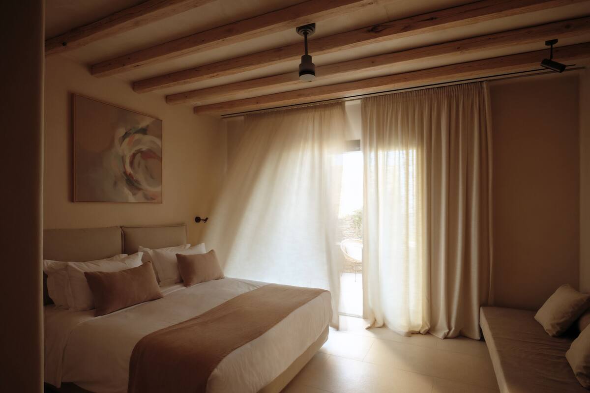 A bedroom with a large bed, a smaller sofa, wooden beams on the ceiling, and a painting on the wall.