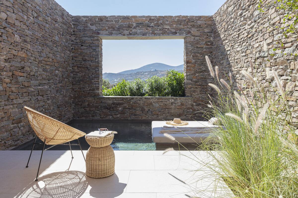 A stone-walled outdoor area with a pool, a wicker chair, a small table, and a view of greenery and mountains.