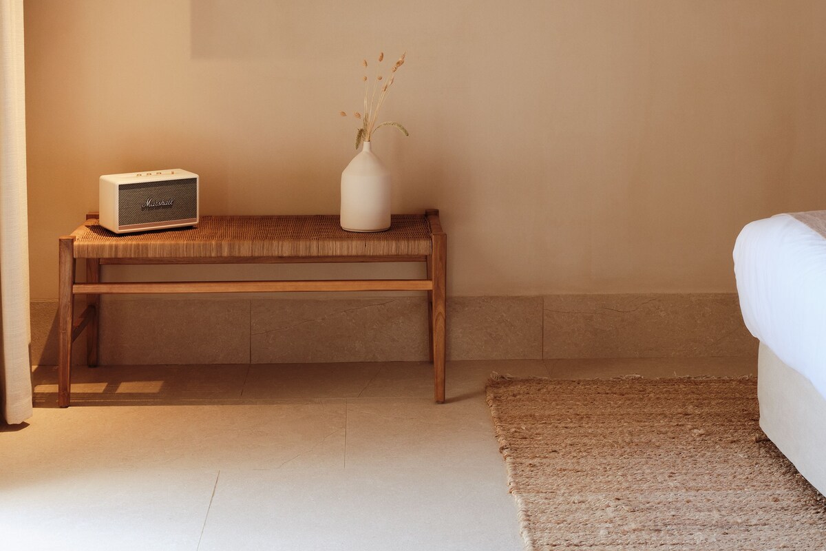 A room with a wooden bench, a white vase with dried plants, and a framed painting on the wall.