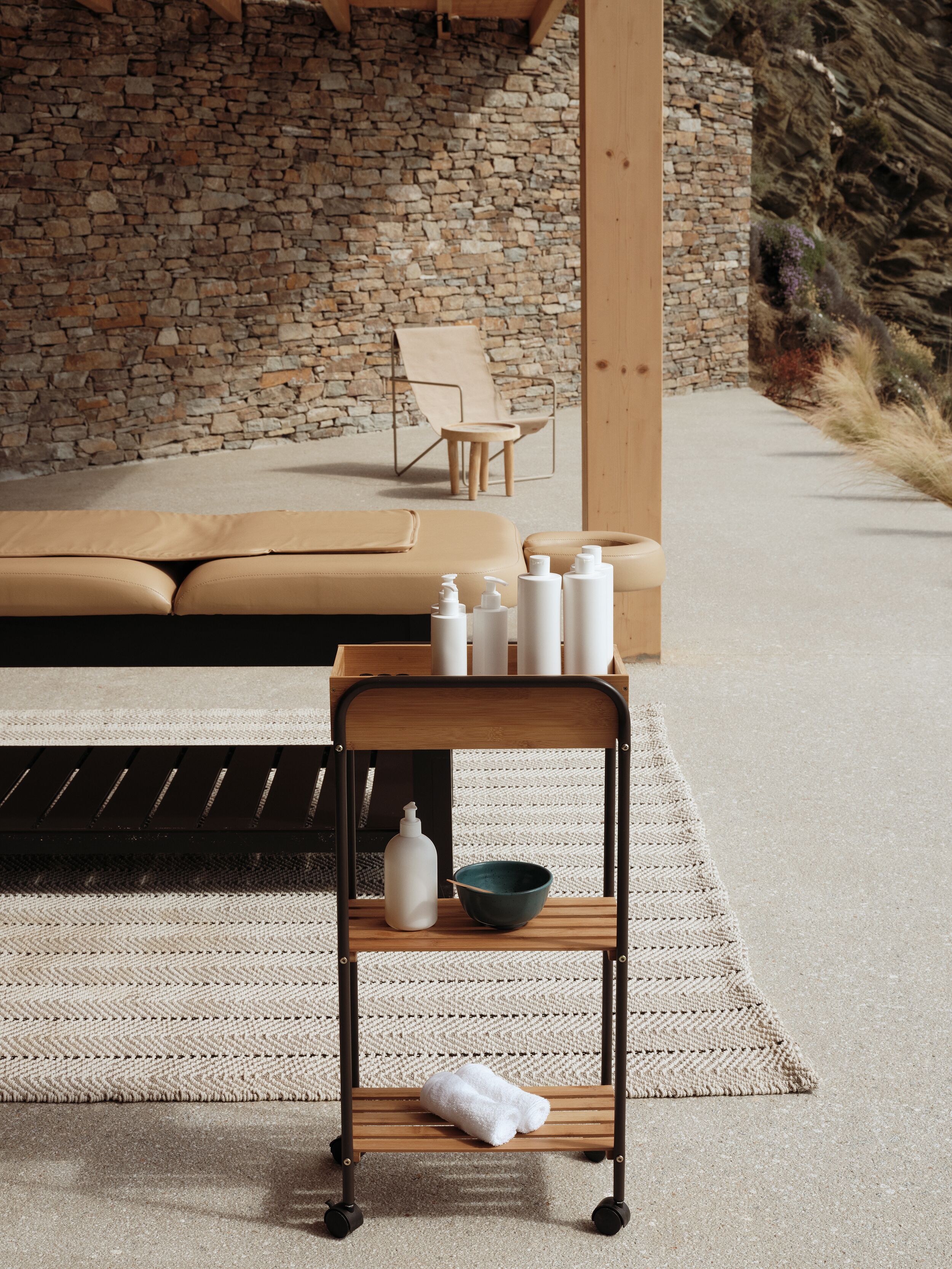 A modern outdoor seating area with a stone wall backdrop, a wooden bench with toiletries, and a lounge chair.