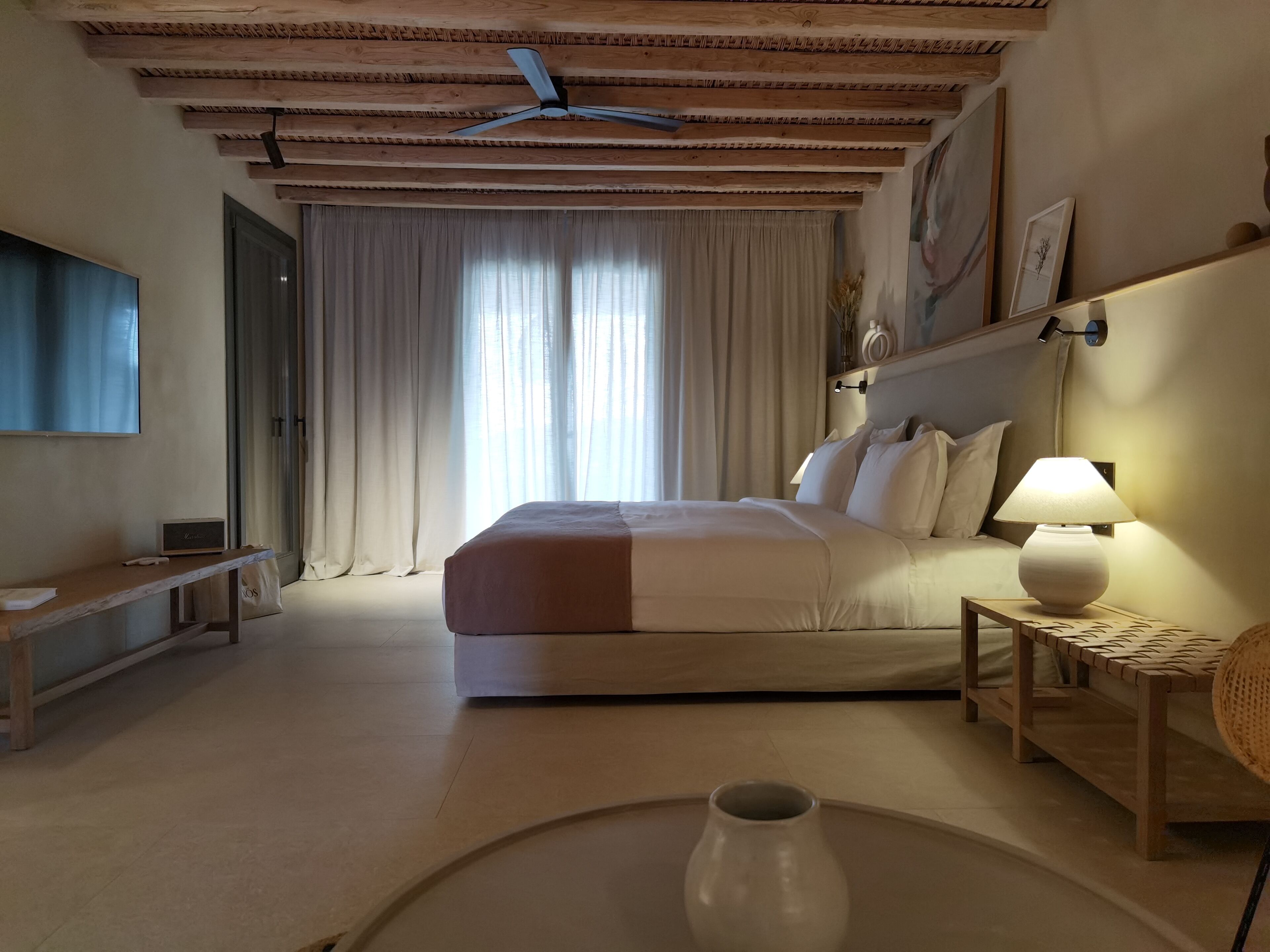 Un dormitorio con cama, lámpara de noche, cabecero de madera y un ventanal con cortinas.