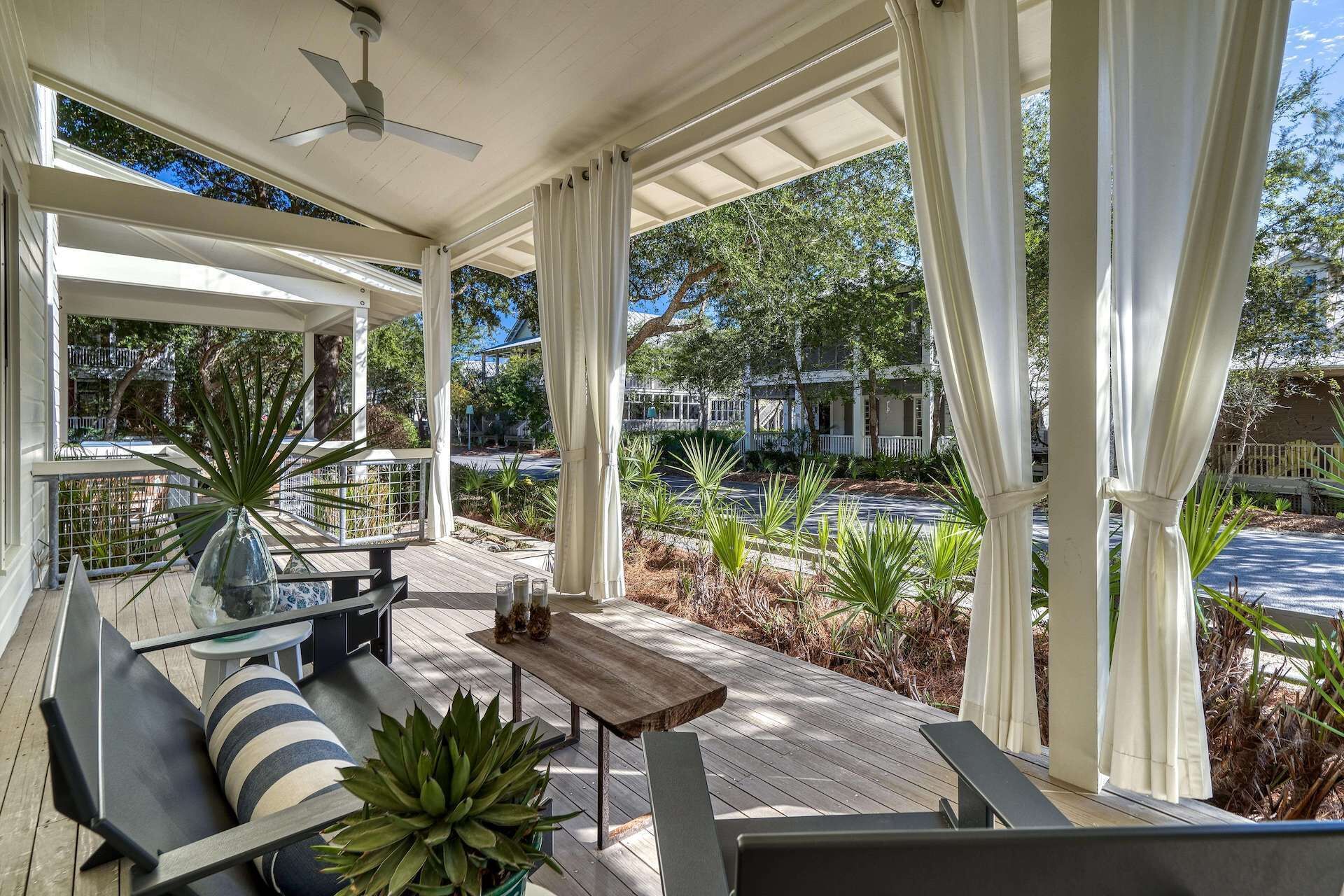 A covered outdoor seating area with a wooden deck, a ceiling fan, and a view of a residential street with houses and trees.