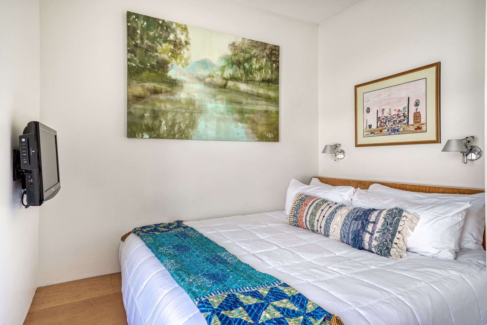 A bedroom with a bed, a painting of a landscape, a framed picture, and a television on the wall.