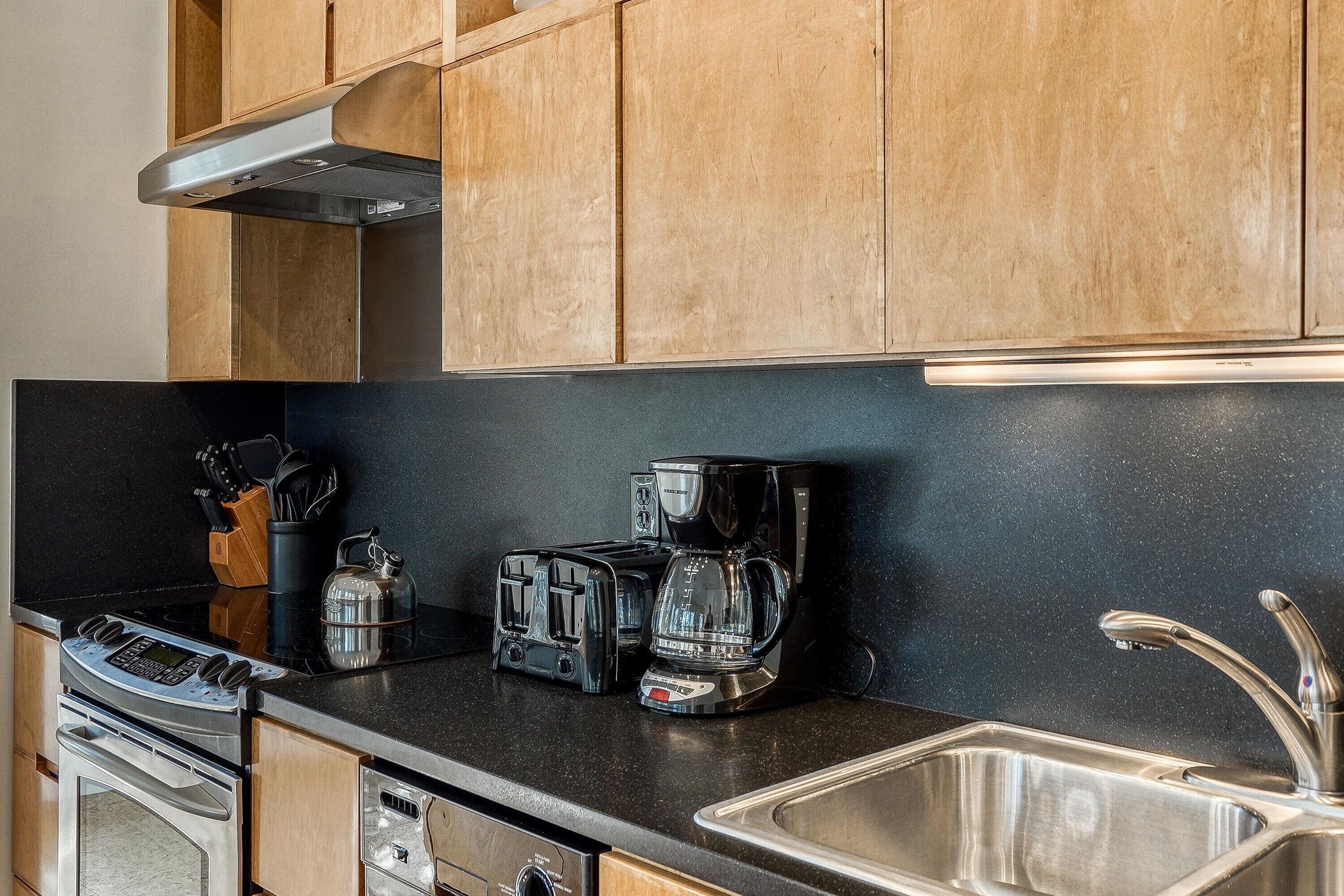 A kitchen with wooden cabinets, a black countertop, a stainless steel sink, a stove, a toaster, and a coffee maker.