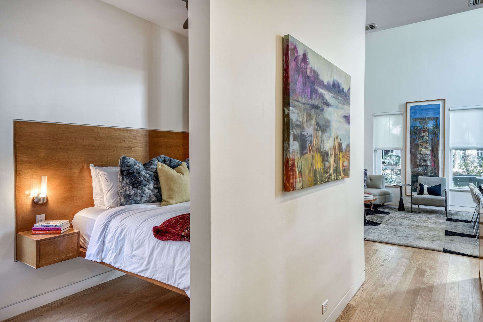 A modern bedroom with a wooden headboard, a bedside table with books, and a large abstract painting on the wall.