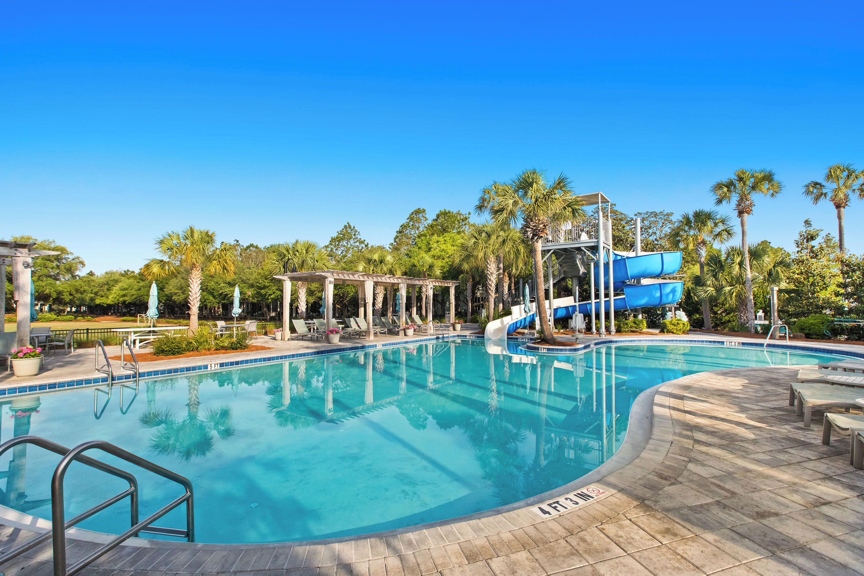 A swimming pool with a water slide, lounge chairs, and palm trees.