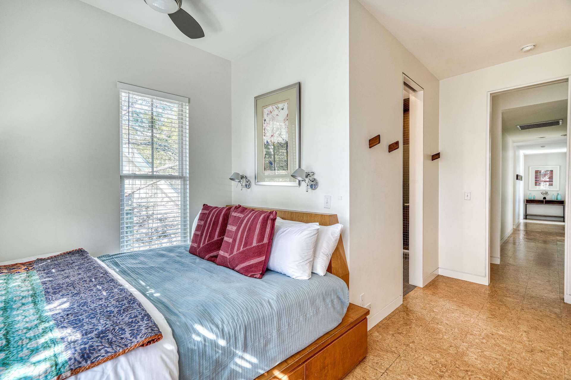 A bedroom with a bed, a window with blinds, a ceiling fan, and a hallway leading to another room.