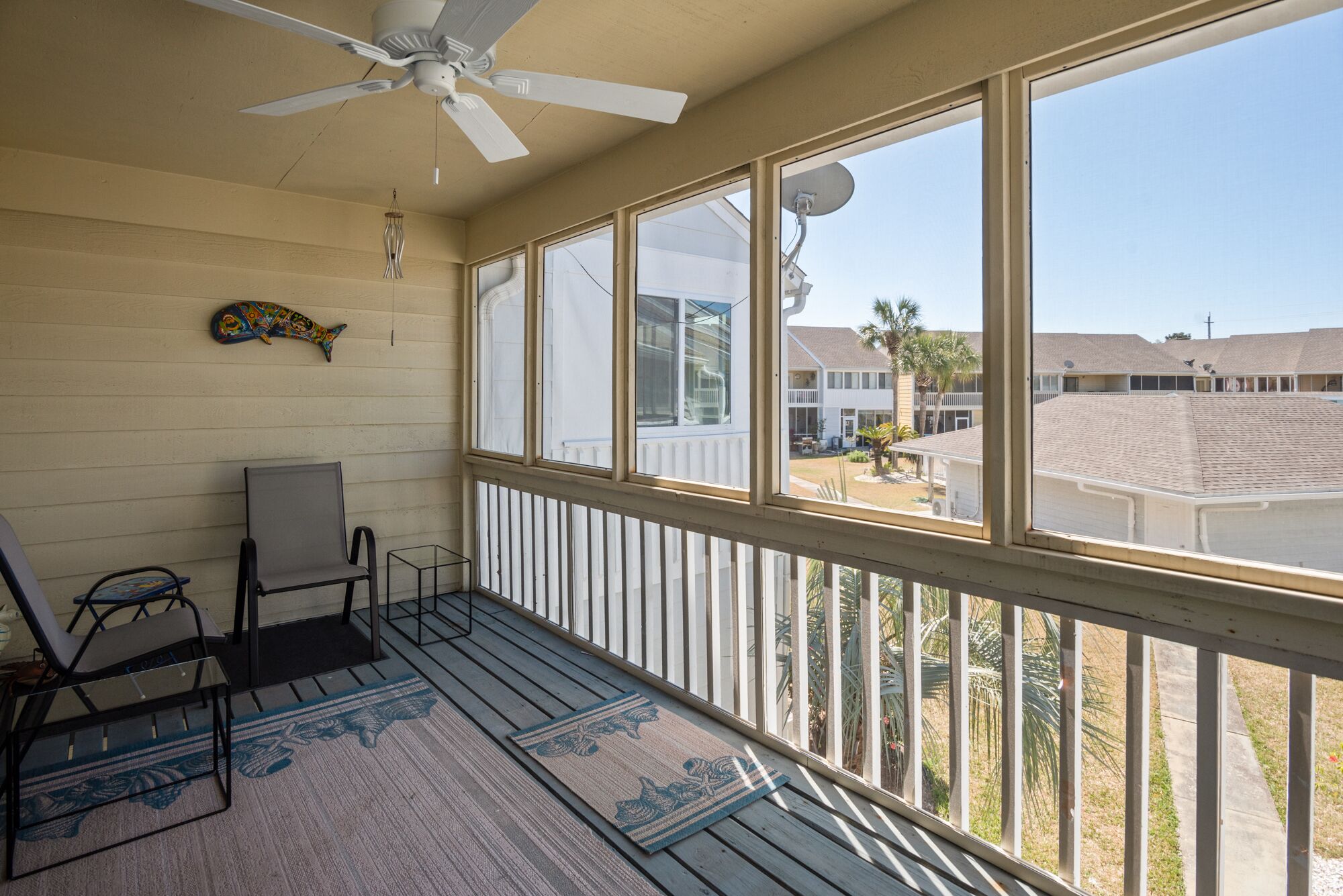 A balcony with a ceiling fan, a chair, a small table, and a view of a residential area with palm trees and houses.
