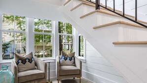 A modern interior with a staircase, two armchairs with patterned cushions, and large windows offering a view of greenery outside. - Amenity Loaded Designer Home on Western Lake with Pool, Elevator, Bikes, LSV (Santa Rosa Beach)