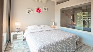 A bedroom with a bed, a nightstand with a lamp, and a window with a view of greenery. - JUNGLE LUXURY FLAT WITH BALCONY AND PRIVATE PARKING IN CASSIS CENTER+WIFI (Cassis)
