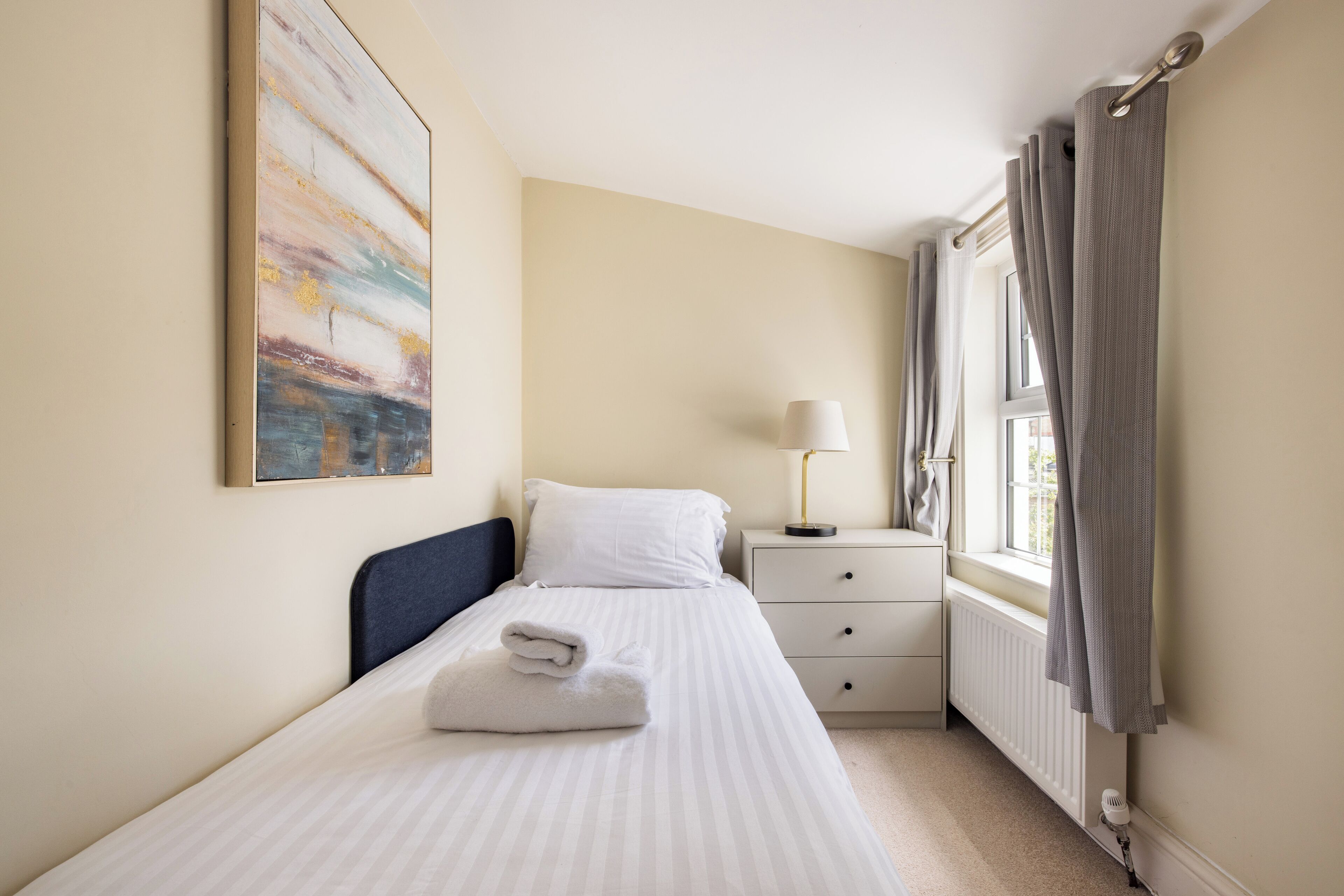 A neatly made bed with a folded towel, a bedside lamp, a window with curtains, and a radiator.