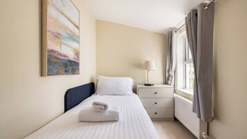A neatly made bed with a folded towel, a bedside lamp, a window with curtains, and a radiator.