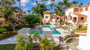 A pool area with a thatched-roof structure, surrounded by palm trees and a white building with red-tiled roofs. - Stunning Ocean View Condo in Punta Cana (Punta Cana)