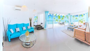 A modern living room with a blue sofa, a glass coffee table, and a pink sofa. There is a large window with a view of palm trees and the ocean. - Stunning Ocean View Condo in Punta Cana (Punta Cana)