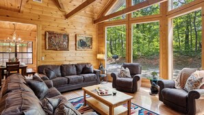A log cabin interior with a large wooden ceiling, a wooden dining table with chairs, a leather sofa, a coffee table, and large windows offering a forest view. - Kozy Bear 3 King BRs | Hot Tub | Pool Table (Sevierville)