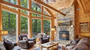 A log cabin interior with a stone fireplace, leather sofas, and large windows overlooking a forest. - Kozy Bear 3 King BRs | Hot Tub | Pool Table (Sevierville)