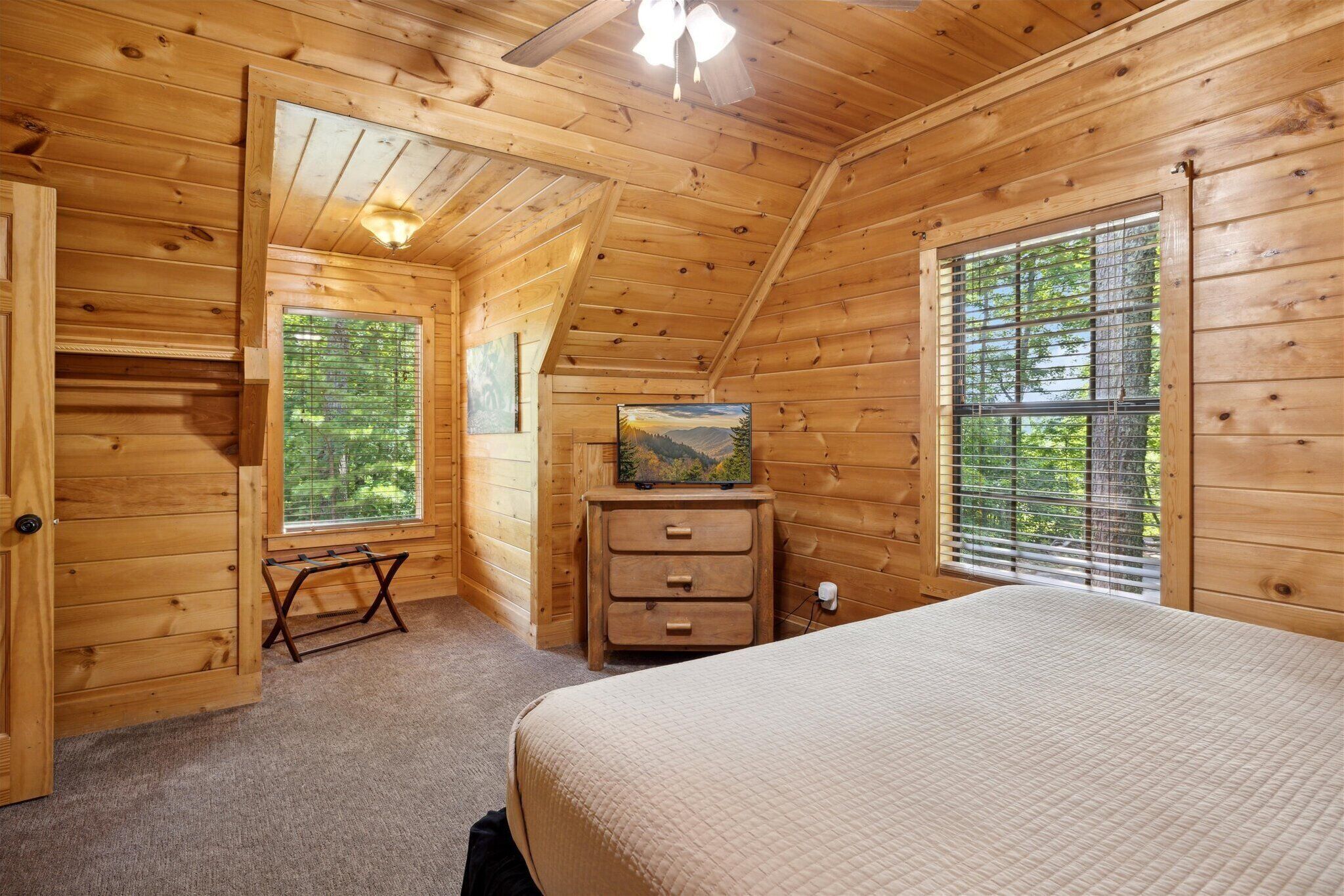 Une chambre dans une cabane en bois, avec un lit, une commode, une fenêtre donnant sur les arbres et un ventilateur de plafond.