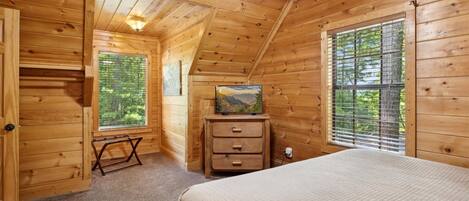 Une chambre dans une cabane en bois, avec un lit, une commode, une fenêtre donnant sur les arbres et un ventilateur de plafond.