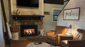 A cozy living room with a stone fireplace, leather sofas, a round ottoman, and a wall-mounted TV.