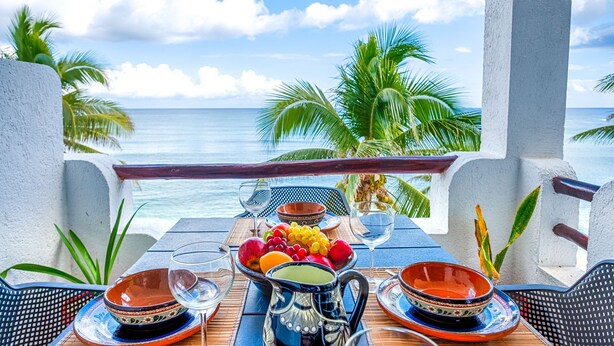 Ein Tisch mit Tellern, Schalen und einer Karaffe, mit Blick auf einen Strand mit Palmen und dem Meer.