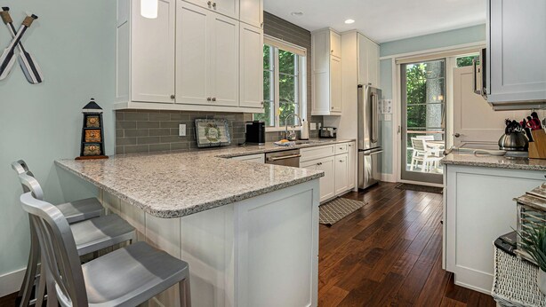 A modern kitchen with white cabinets, granite countertops, and stainless steel appliances. There is a large island with bar stools, and a dining area with a view of the outdoors. - Sleepy Hollow Beach Resort, Cottage 9 – Family Fun on Lake Michigan! (South Haven)
