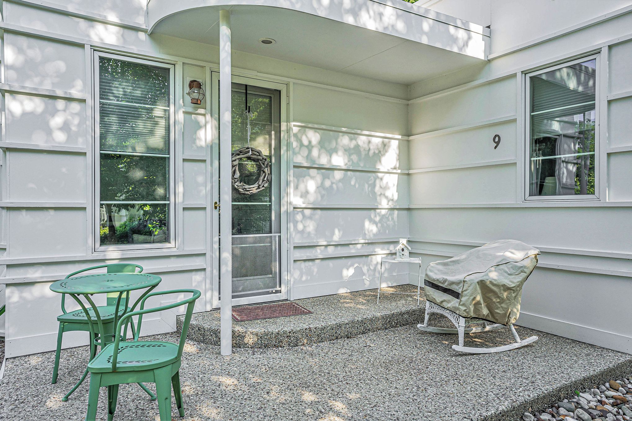 Une façade blanche, une chaise berçante placée sous un auvent, une petite table avec des chaises et une porte décorée d’une guirlande.