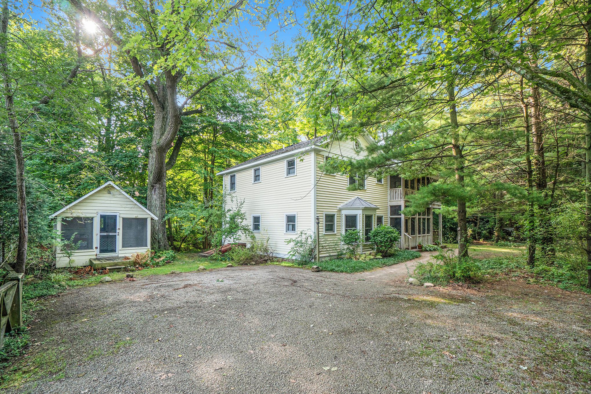 A two-story house with a gravel driveway, surrounded by dense trees and a small shed.