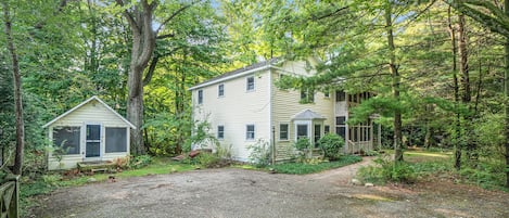 A two-story house with a gravel driveway, surrounded by dense trees and a small shed.