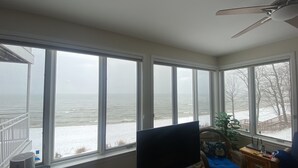 Una habitación con un ventanal grande con vistas a un paisaje nevado, un ventilador de techo y un televisor sobre un soporte.