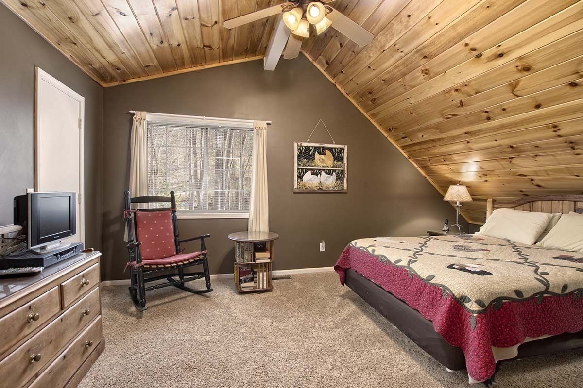 A bedroom with a sloped ceiling, a bed with a patterned blanket, a chair, a small table, and a television on a dresser.