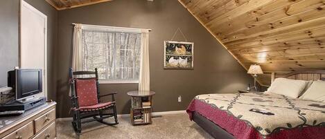 A bedroom with a sloped ceiling, a bed with a patterned blanket, a chair, a small table, and a television on a dresser.