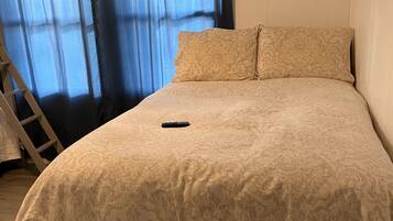 Une chambre à coucher avec un lit, deux oreillers, une télécommande et une fenêtre avec des rideaux.