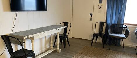 A room with a flat-screen TV mounted on the wall, a wooden table with chairs, a door with a lock, and a window with blue curtains.