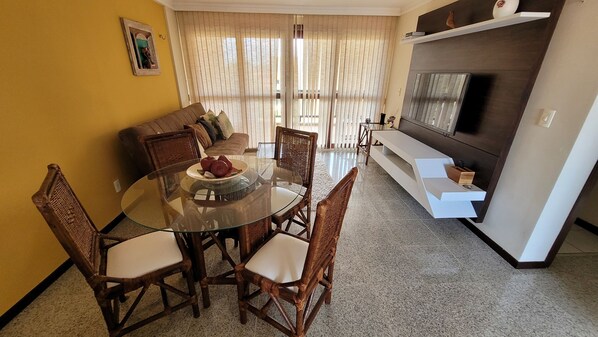 A dining area with a glass table and wicker chairs, a sofa, and a TV on a shelf. - Flat na Praia de Búzios RN por Carpediem (Nisia Floresta)