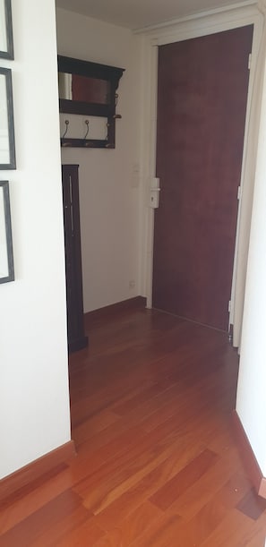 A hallway with wooden flooring, a dark wooden door, and a white wall with a mirror and hooks.