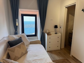 A bedroom with a bed, a nightstand, a lamp, and a view of the sea through a window.