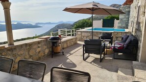 An outdoor patio with a stone wall, a grill, a table, and chairs. There is a pool in the background with a view of the sea and mountains. - Amber's Place, Cosy House With Pool and sea Views (Dubrovnik)