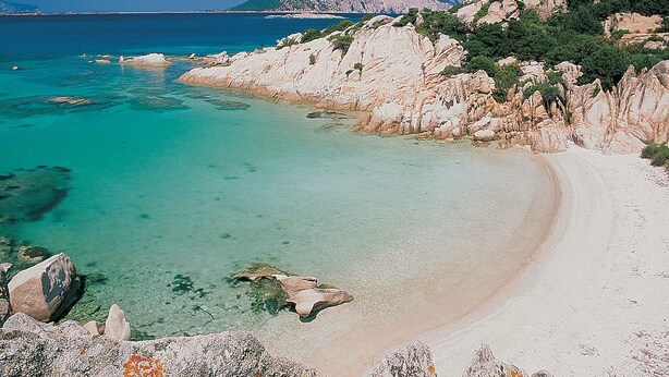 Una playa costera con aguas turquesas cristalinas, acantilados rocosos y una montaña al fondo.