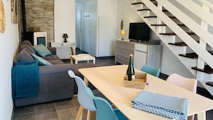 A modern living room with a sofa, dining table, and chairs. A staircase with a white railing leads to another level. - Villa du Pêcheur, Gâvres Peninsula (Gâvres)