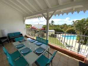 Un área de comedor al aire libre techada con una mesa preparada para cuatro personas, sillas y vistas a una piscina y follaje.