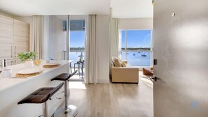 Eine moderne Küche mit einer großen Kochinsel, ein Essbereich mit Barhocker, eine Couch neben einem großen Fenster mit Blick auf ein Gewässer und eine Aussicht auf Boote.