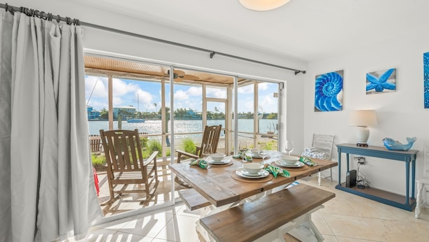 Uma área de refeições com vista para o mar, uma mesa de madeira posta com pratos e copos, e um objeto decorativo azul em um console.