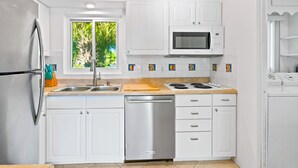Uma cozinha moderna com armários brancos, geladeira de aço inoxidável, micro-ondas, lava-louças e uma janela com vista para o verde.