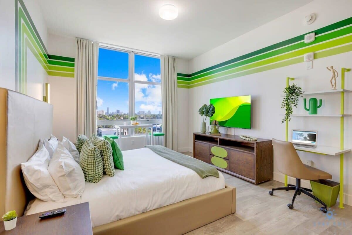 A modern hotel room with a large bed, a TV, a desk with a chair, and a view of the city.