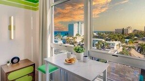 Ein modernes Hotelzimmer mit großem Fenster und Blick auf die Stadt, ein kleiner Tisch mit einem Teller Croissants sowie ein Nachttisch mit einer Uhr und einer Pflanze.
