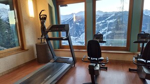 Ein Fitnessraum mit Laufband, Trainingsstühlen und Blick auf verschneite Berge durch große Fenster.