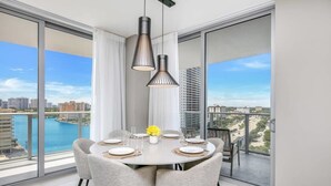 A dining area with a round table set for four, large windows offering a view of a cityscape and water, and modern pendant lighting.