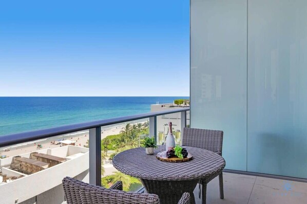 A balcony with a wicker table set with a bottle, glasses, and grapes, overlooking a beach and ocean.